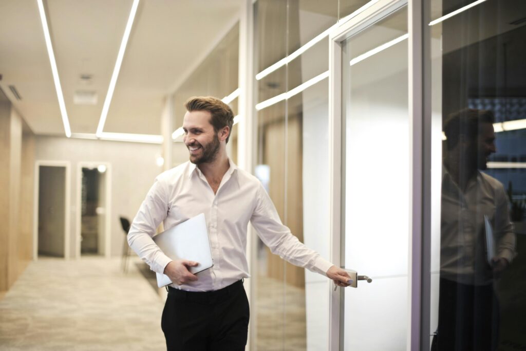 A smiling man holds a laptop while standing in a modern office hallway, exuding confidence.